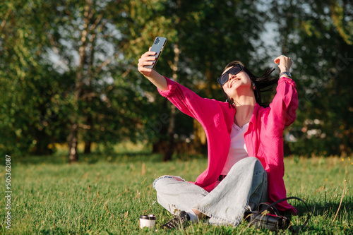 Happy young woman in sunglasses with cellphone sitting on grass at the park in sunny day. Woman takes picture of herself