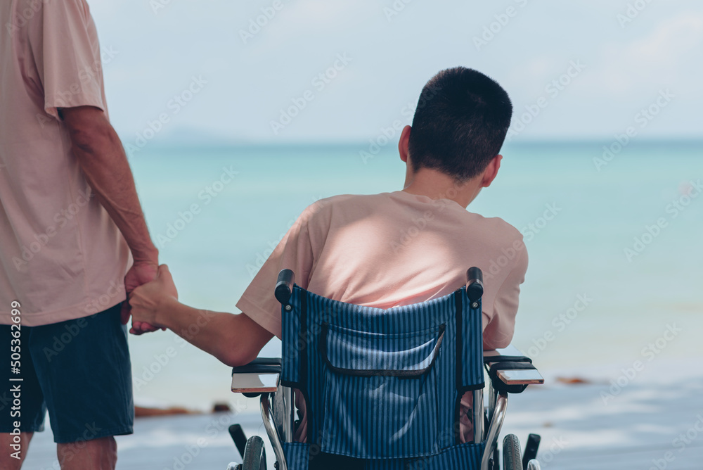 Φωτογραφία Wheelchair and behind of young man and father's, Dad hand holding the son's hand staring at the sea ahead,Family vacation activity,Encourage to strengthen,Mental health and travel or vacation concept