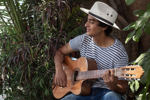 young hispanic musician enjoys playing the acoustic guitar outdoors among the plants in the garden, he enjoys his hobby very much and the expression on his face confirms it.