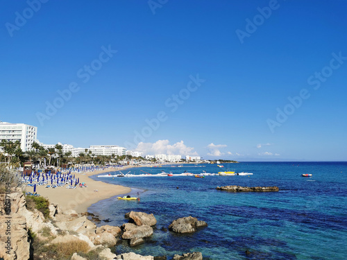 Large stones near the shore, clear water of the Mediterranean Sea, a view of the sandy large Sunrise beach and hotels against the background of a blue sky with clouds.