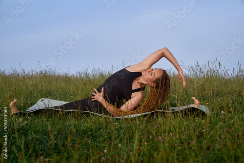 Front view of bloody smiling girl doing side banding on yoga mat among grass. Stretching of beautiful woman, deeply breathing. Concept of yoga outdoors.