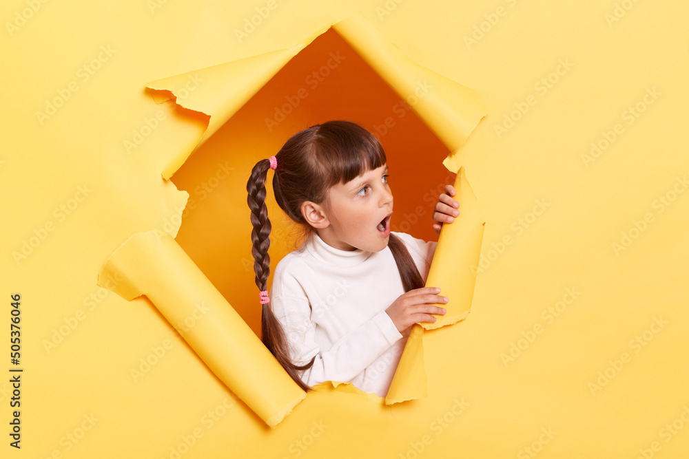 Φωτογραφία Shocked surprised little girl with braids wearing casual shirt looking through torn hole in yellow paper, looking aside with open mouth, sees shocking advertisement