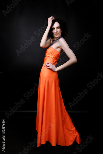 Brunette girl in an orange long dress posing in a photo studio on a dark background