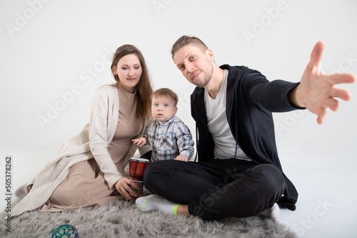 Fototapeta Naklejka Na Ścianę i Meble -  Young caucasian parents with one year old son sit on carpet in white studio.