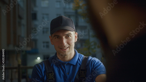 Young delivery man courier in blue cap smiling into customer takes order around