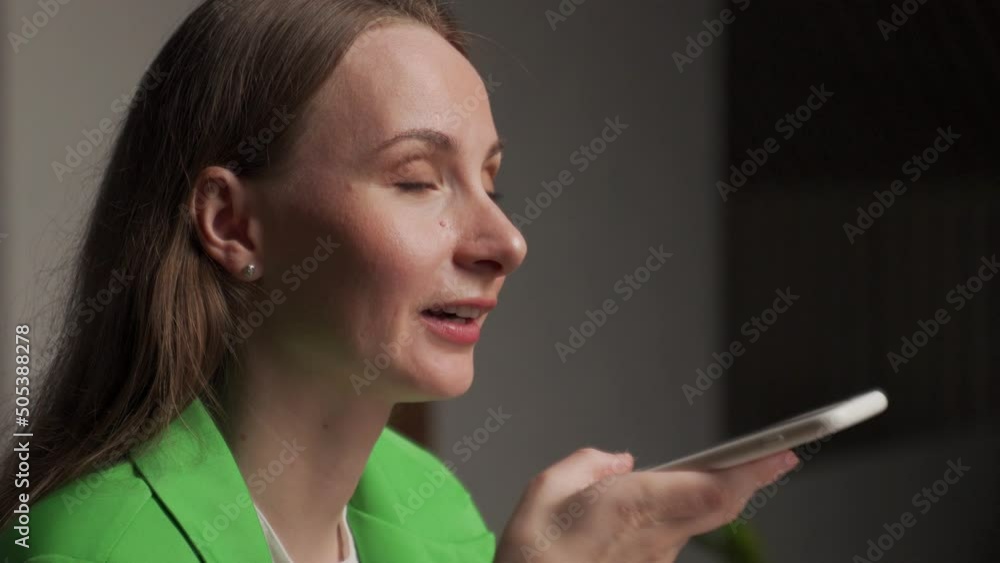 Young woman calls to friend via mobile phone telling about first working day. Brunette lady in green jacket holds device on blurred background closeup Young woman calls to friend via mobile phone telling about first working day. Brunette lady in green jacket holds device on blurred background closeup