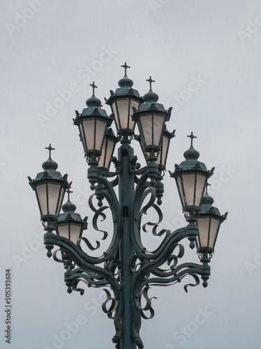 Old fashioned lamp. Vintage street lamp on a blue sky background. Street lamp on the background of the blue sky. Antique wrought iron lantern with glass inserts on the background of the blue sky. © Dmitry Presnyakov