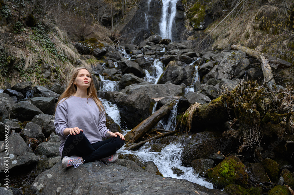 Girl sits on a large stone and meditates near a waterfall next to a mountain river. Girl sits on a large stone and meditates near a waterfall next to a mountain river.
