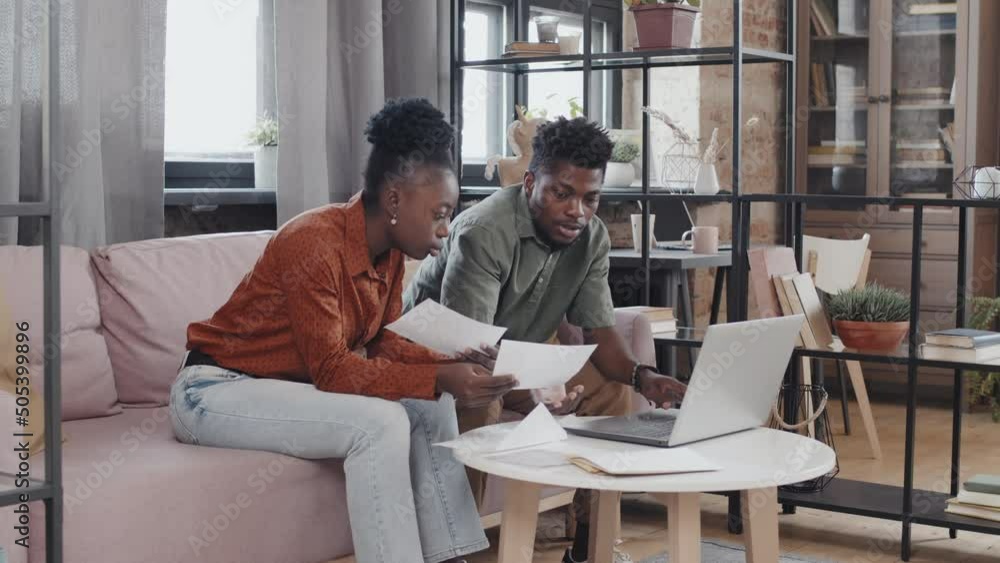 Medium long of young African American woman and man sitting on couch in living room, doing paperwork, using portable computer and having conversation Medium long of young African American woman and man sitting on couch in living room, doing paperwork, using portable computer and having conversation
