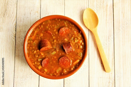 Clay bowl of lentils soup with Spanish sausage or chorizo with wooden spoon beside the bowl, traditional food of Spain. photo isolate on wooden background top view