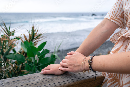 Close-up of white-skinned female hands resting on a wooden railing in front of the sea. Horizontal format