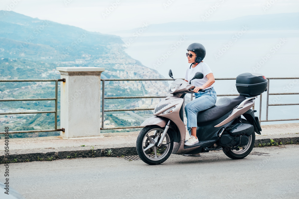 Obraz premium Middle-age woman in helmet and sunglasses on motor scooter on the Sicilian old town streets in the Forza d'Agro with Sant'Alessio Siculo harbor. Happy Italian vacation and transportation concept.