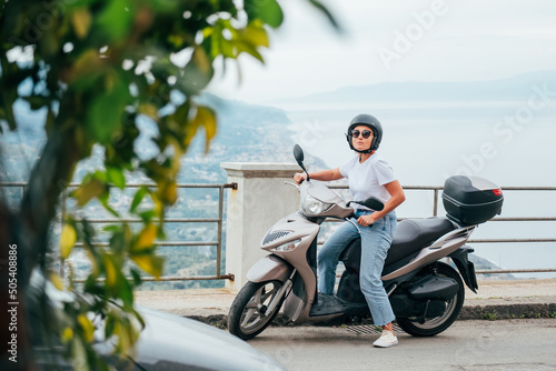 Middle-age woman in helmet and sunglasses on motor scooter on the Sicilian old town streets in the Forza d Agro with Sant Alessio Siculo harbor. Happy Italian vacation and transportation concept.