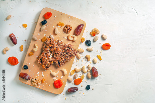 granola bars, dried fruits and nuts lie on top of each other on a wooden board with dried fruits, cereals and nuts scattered nearby, Flatley, kopispace. the board lies diagonally.