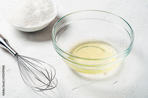 ingredients for the French meringue dessert are egg white, sugar and a whisk on a gray surface, close-up.