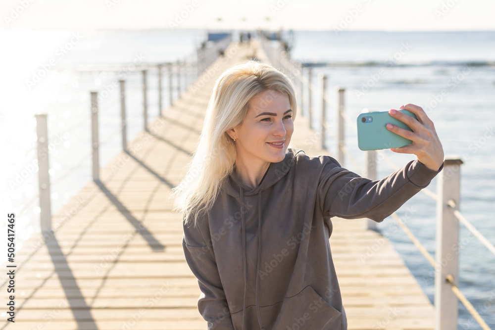 pretty young woman in hoodie uses mobile phone for communication poses, enjoys spending summer vacation at sea tourist town chats online.