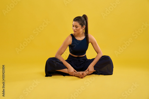 Pretty girl sitting in butterfly II yoga pose, with closed eyes and turned head while exercising. Front view of barefoot woman practicing yoga, isolated on orange studio background. Concept of yoga. © serhiibobyk