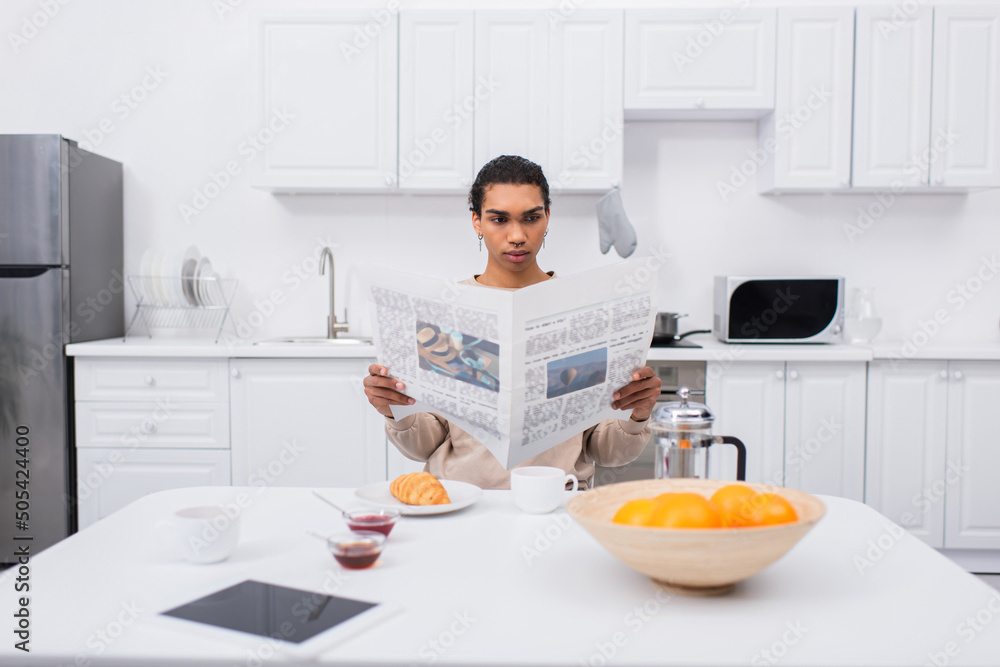 african american man reading newspaper near morning breakfast and digital tablet on table. african american man reading newspaper near morning breakfast and digital tablet on table.