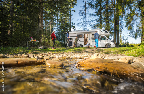 Five Caucasian People on Their Motorhome Road Trip Camping. Riverside RV Dry Campsite with Friends. Hanging Next to Campfire.