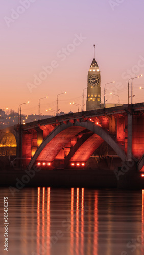 Krasnoyarsk Clock Tower is located in the center of Krasnoyarsk city in Russia against the background of a communal bridge with a beautiful red backlight the enisey river photo