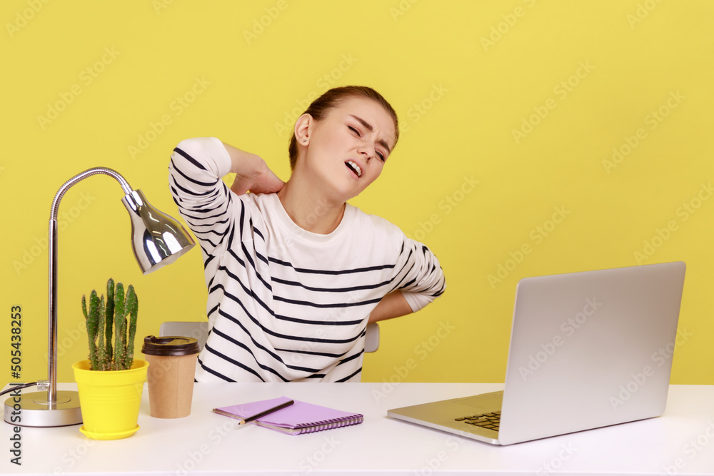 Φωτογραφία Exhausted woman office worker wearing striped shirt sitting on workplace and suffering terrible pain in neck and back, looks tired