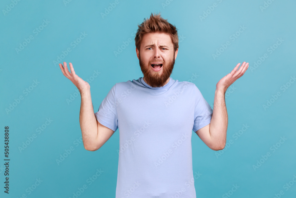 What do you want? Portrait of annoyed frustrated bearded man standing with raised hands and indignant face asking why, annoyed by problem. Indoor studio shot isolated on blue background. What do you want? Portrait of annoyed frustrated bearded man standing with raised hands and indignant face asking why, annoyed by problem. Indoor studio shot isolated on blue background.