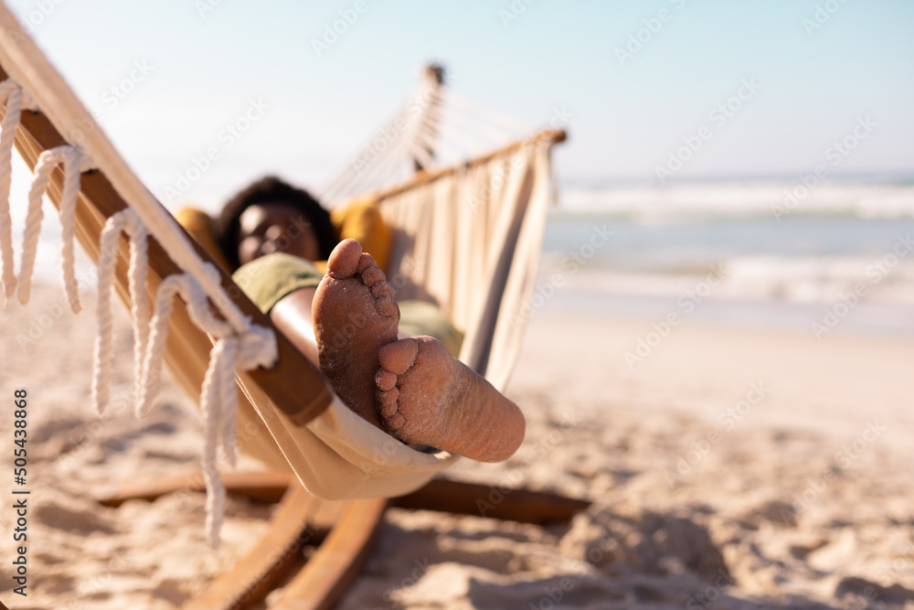 Obraz premium African american mature woman with sand on feet lying on hammock against sea and sky in summer