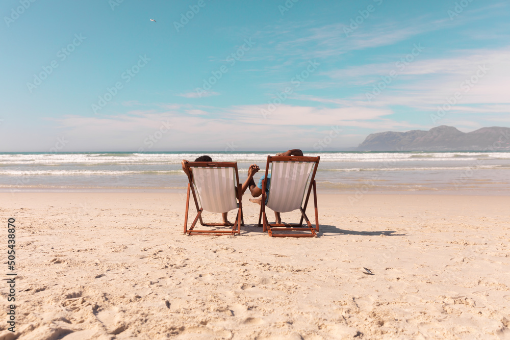African american couple holding hands while relaxing on deckchairs at beach against sky in summer - obrazy, fototapety, plakaty 