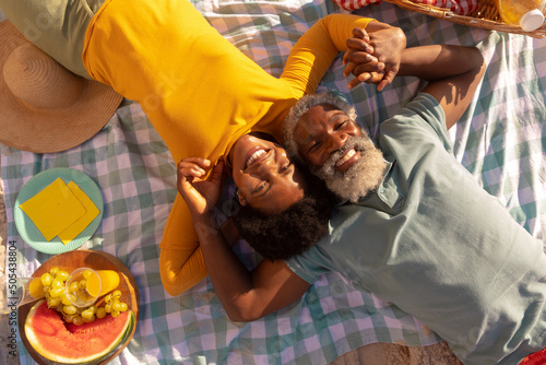 Overhead view of happy african american couple holding hands by food while lying on blanket at beach