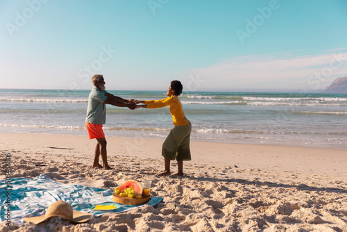 African american couple holding hands and dancing on sandy beach against sea and clear sky in summer