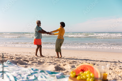 African american couple holding hands and dancing on sandy beach against sea and blue sky in summer