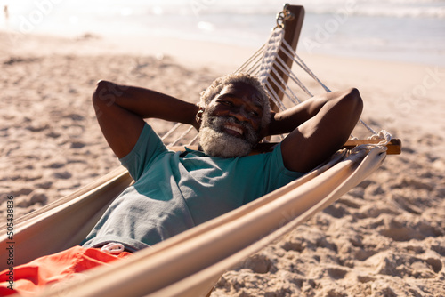 Bearded african american senior man with hands behind head lying on hammock at beach in summer
