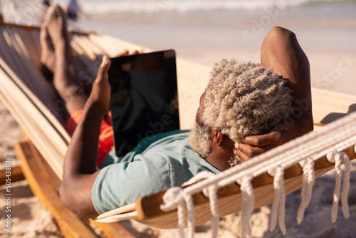 African american senior man with gray hair using digital tablet while lying on hammock at beach