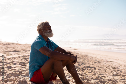 Thoughtful bearded african american senior man with gray hair sitting on sandy beach against sky
