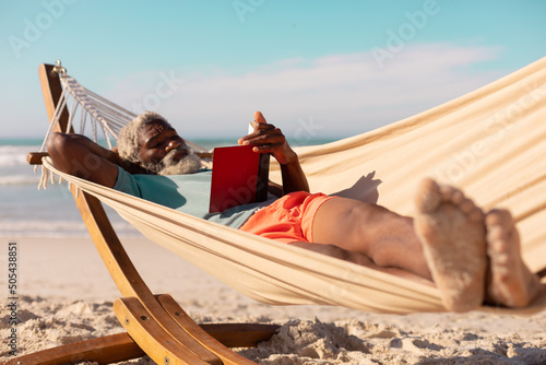 African american bearded senior man reading book while lying on hammock at beach against sky