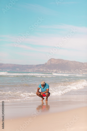 African american senior man with gray hair crouching at shore against blue sky on sunny day