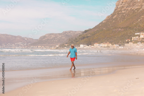 Full length of african american senior man walking at shore against mountain and sky in summer