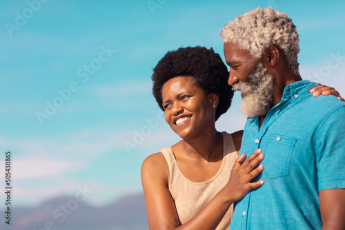 Bearded african american senior man and happy mature woman with afro hair at beach against blue sky
