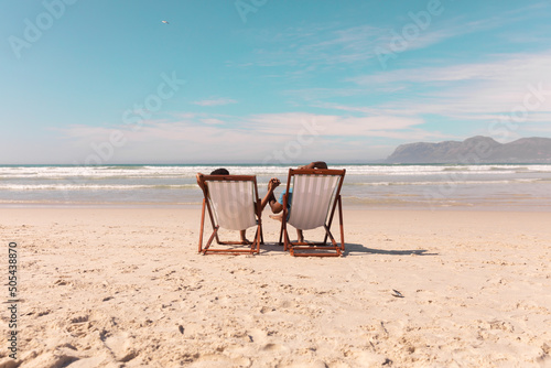 African american couple holding hands while relaxing on deckchairs at beach against sky in summer