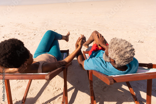 High angle view of african american couple holding hands while sitting on deckchairs at sandy beach