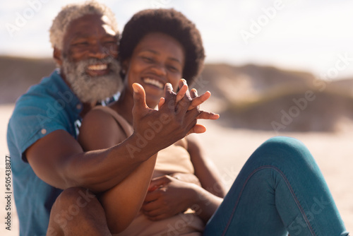 Romantic happy african american couple holding hands while sitting at beach against sky in summer