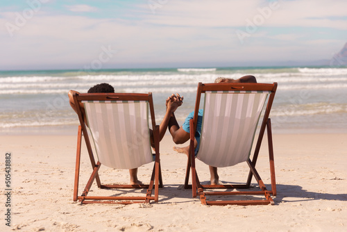 African american couple holding hands while relaxing on deckchairs at beach against cloudy sky