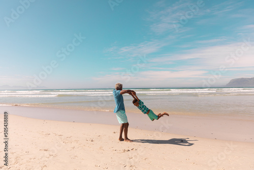 African american senior man holding grandboy's hands and spinning at beach against sky in summer