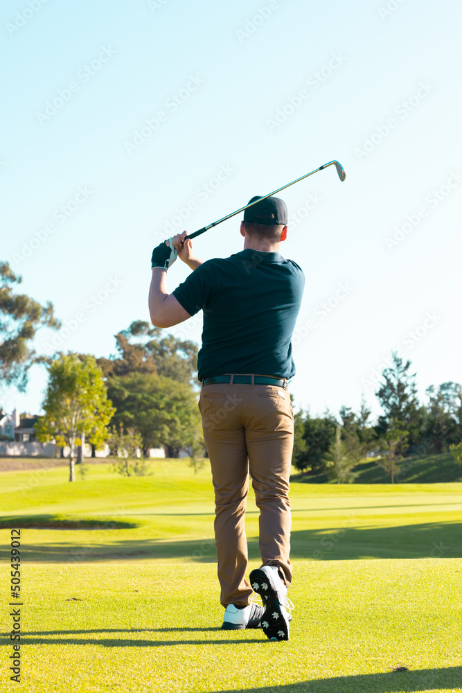 Obraz premium Rear view of caucasian young man hitting with golf club against clear sky at golf course in summer