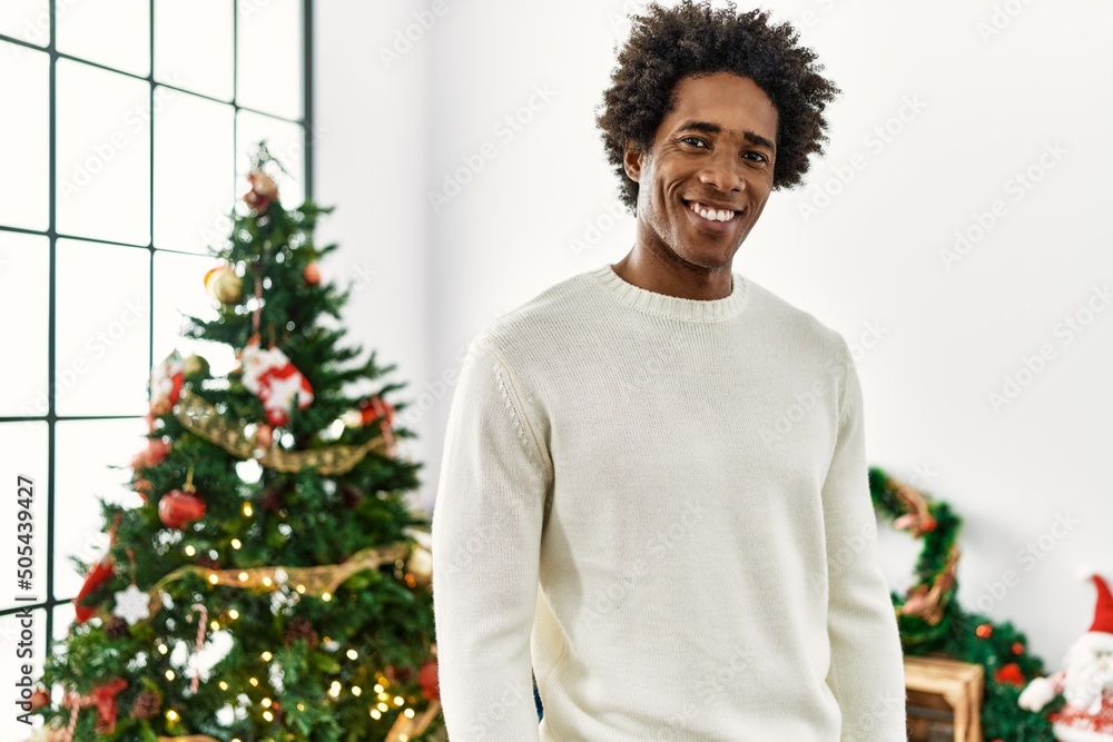Young african american man smiling happy standing by christmas tree at home. Young african american man smiling happy standing by christmas tree at home.