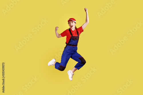 Fototapeta Naklejka Na Ścianę i Meble -  Full length portrait of extremely happy worker woman jumping and clenched fists, celebrating long awaited day off, wearing overalls and red cap. Indoor studio shot isolated on yellow background.