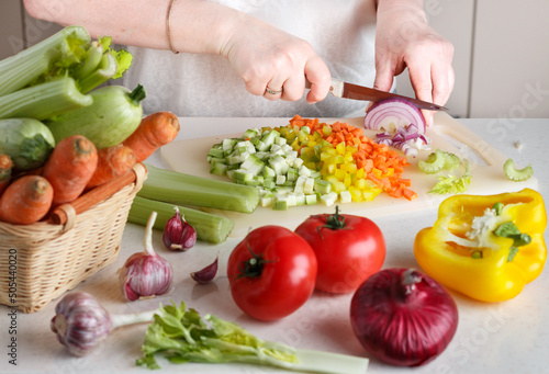 Women's hands chop fresh vegetables for cooking. Carrots, zucchini, celery, bell pepper, tomatoes, garlic and red onion on a cutting board. Culinary ingredients. Healthy eating. Diet. Selective focus