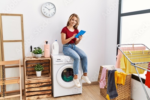 Young caucasian girl using touchpad waiting for laundry sitting on whasing machine at home. photo