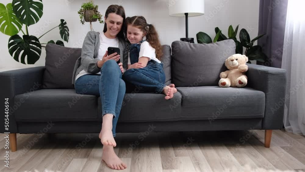 Happy mum and cute kid daughter having fun using cellphone. Young nanny playing mobile games with adorable small child girl, resting on cozy sofa in living room at home. Addicted to modern technology Happy mum and cute kid daughter having fun using cellphone. Young nanny playing mobile games with adorable small child girl, resting on cozy sofa in living room at home. Addicted to modern technology