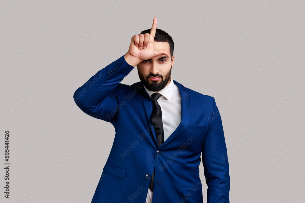 Φωτογραφία Portrait of upset bearded man showing loser gesture, L finger sign on forehead, upset about dismissal, unlucky day, wearing official style suit